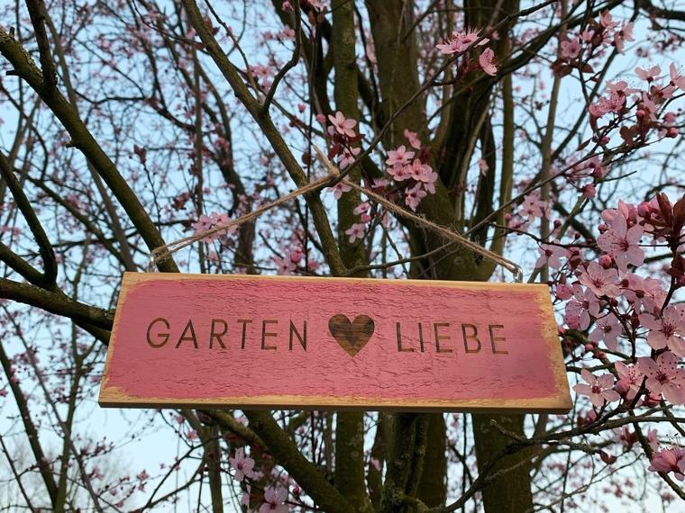 Beerenrosa gestrichenes Gartenschild mit dem Schriftzug Garten Liebe vor dunkler Baumrinde